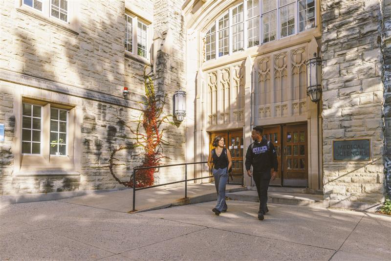 Two students walk and converse outside the entrance of the Medical Sciences building, surrounded by historic stone architecture and autumn foliage